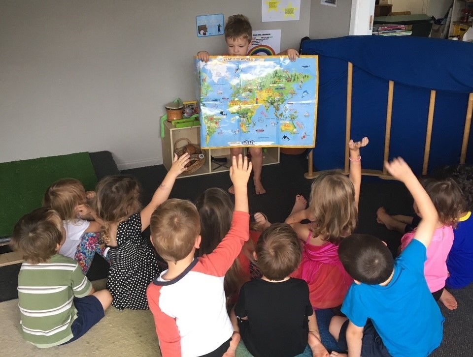 Children raising their hands in class while looking at their teacher holding a world map