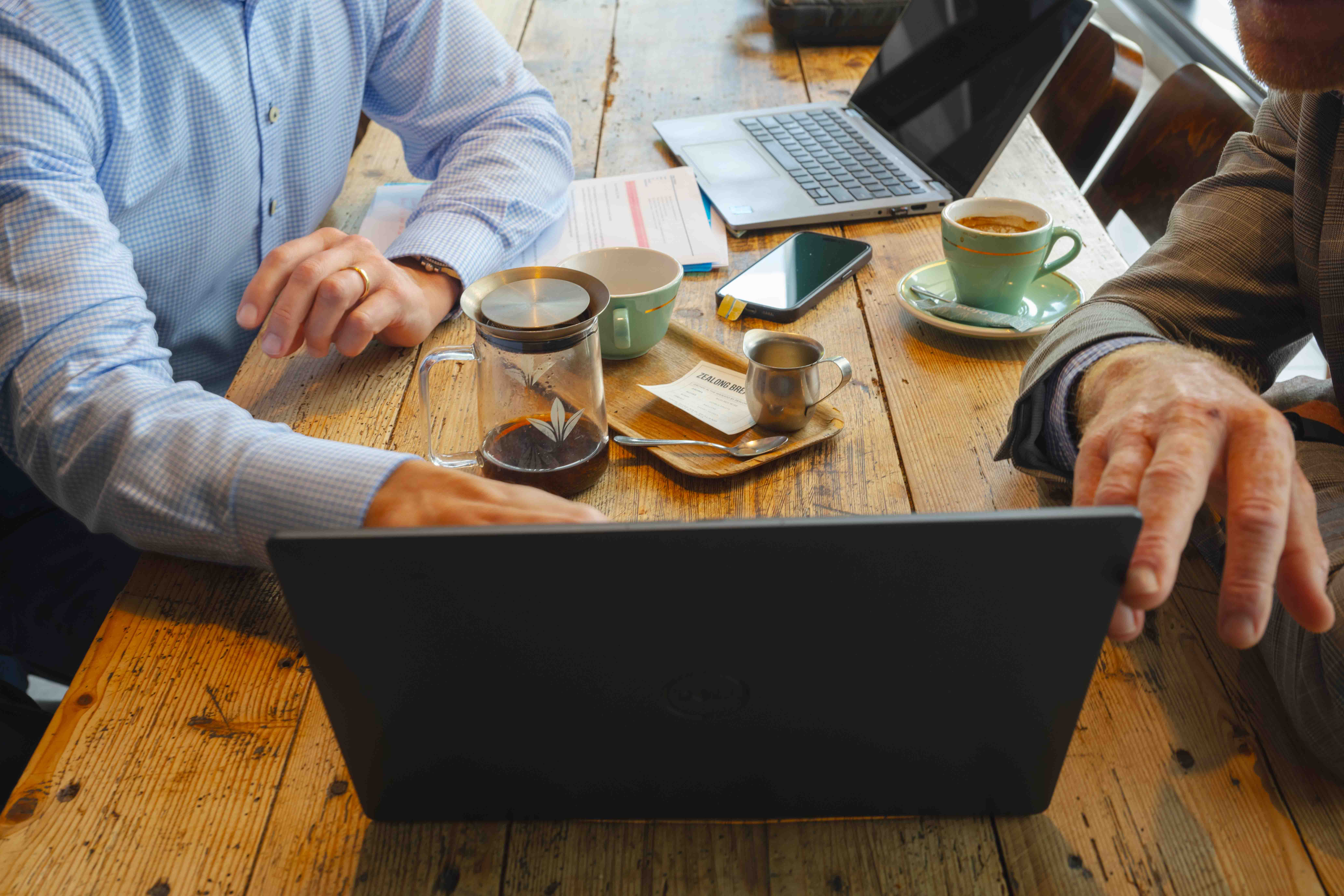 Close up of two people sitting at a table and pointing at a laptop, with tea and cups on the table.