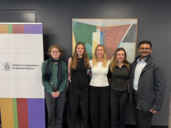 Group portrait of the five interns with a colourful piece of artwork in the background, and a banner with the Ministry logo to the left of the group.