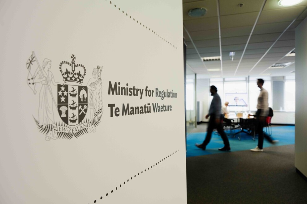 Two people walking in an office, with the Ministry's logo on a white banner in the foreground.